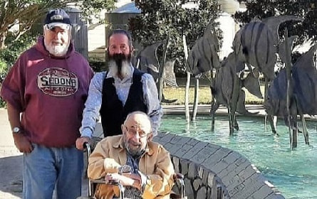 Portrait of 3 men outside the entrance to the Manteo Aquarium in NC. 2 men by a fountain with fish decor and one man in a wheelchair