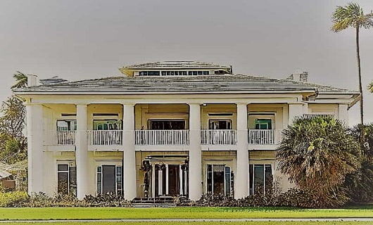 Southern style home with palms planted on the grounds. White columned two-story home with palm trees and a green front lawn.