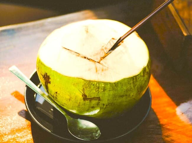 Split fresh coconut on a plate on a table with a spoon nearby and a straw to drink the fruit water.